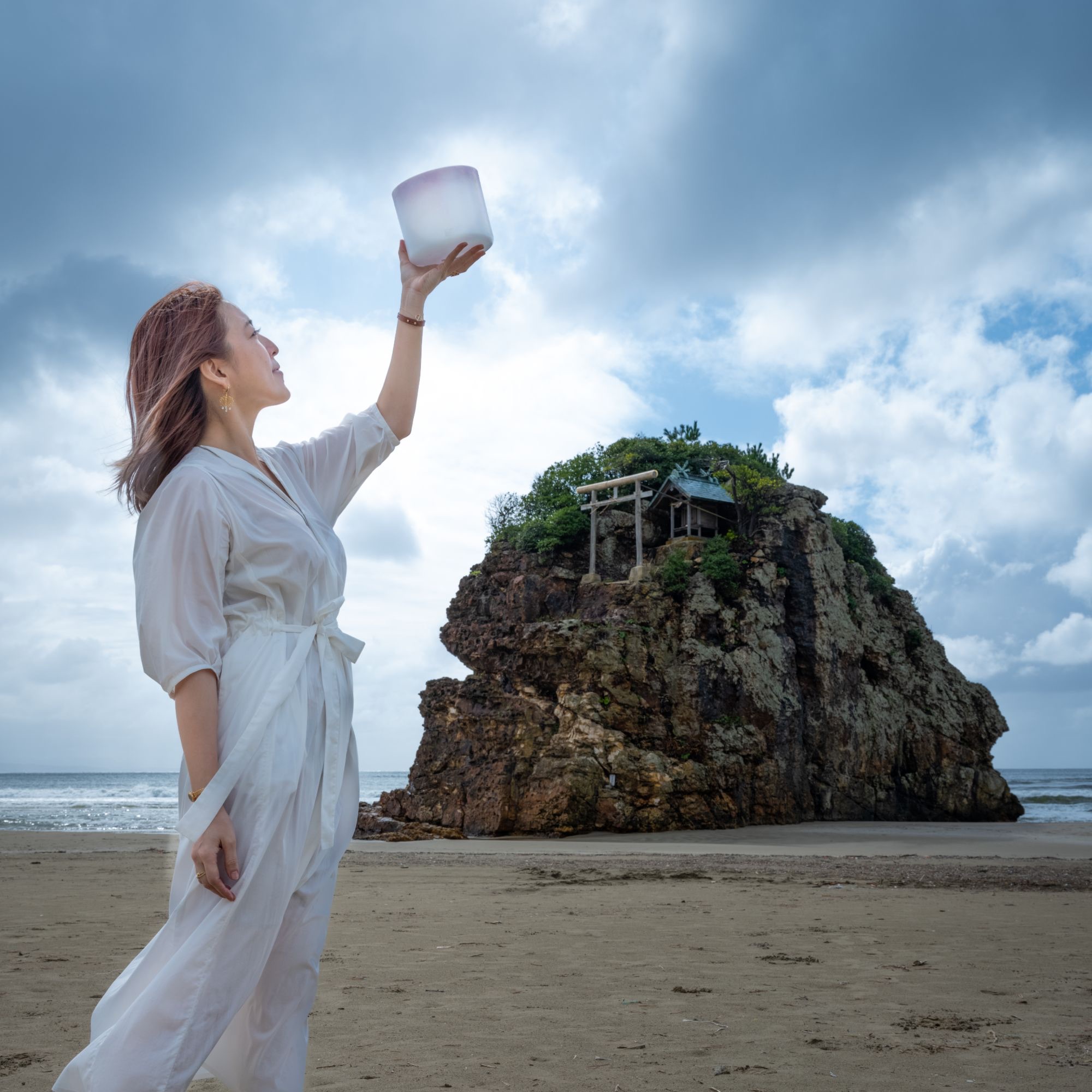 稲佐の浜で、弁天島を背に空へクリスタルボウルを掲げる mogmin.